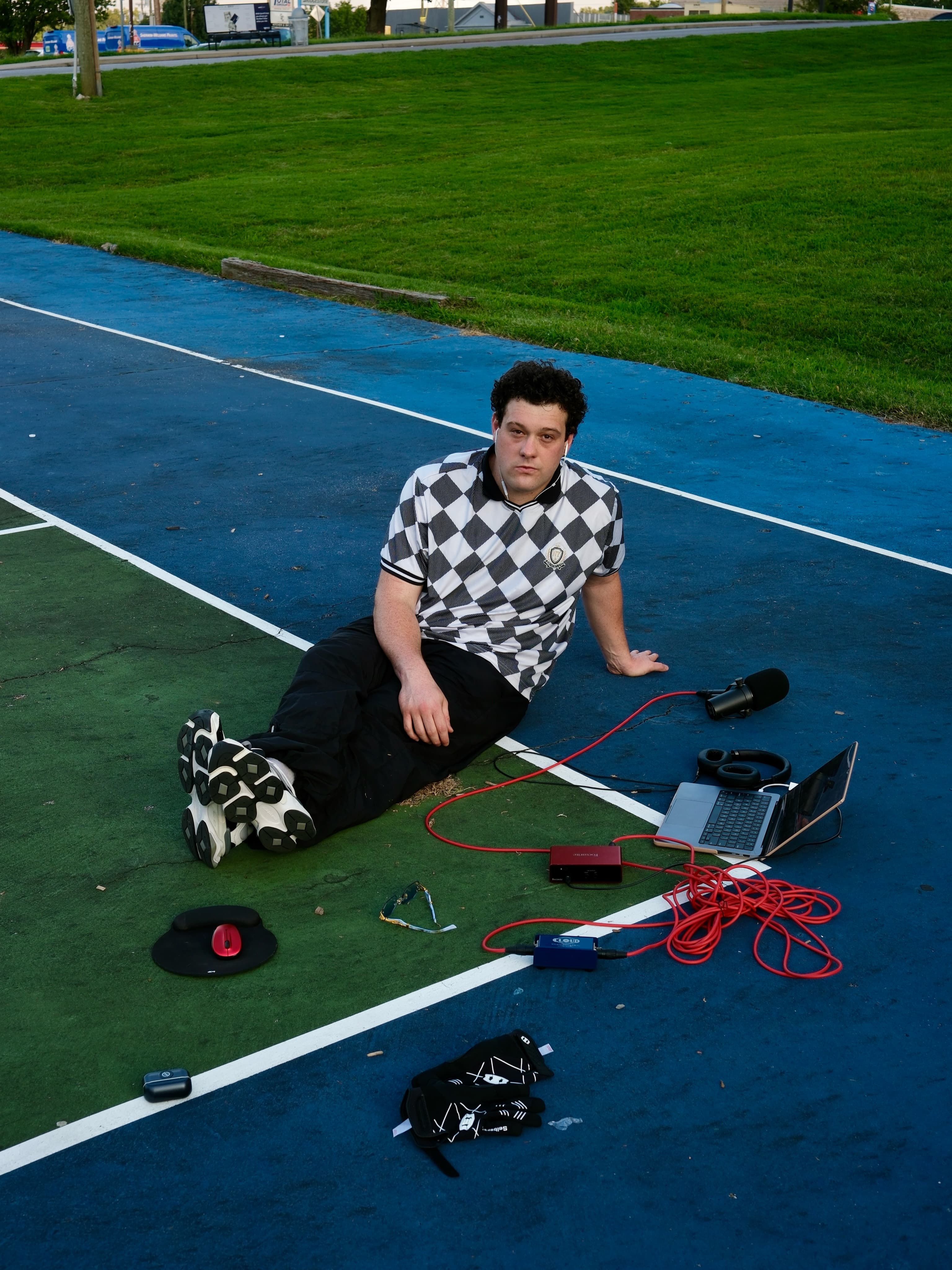 BRAVEN looking up at the camera, seated beside his recording setup on the court.