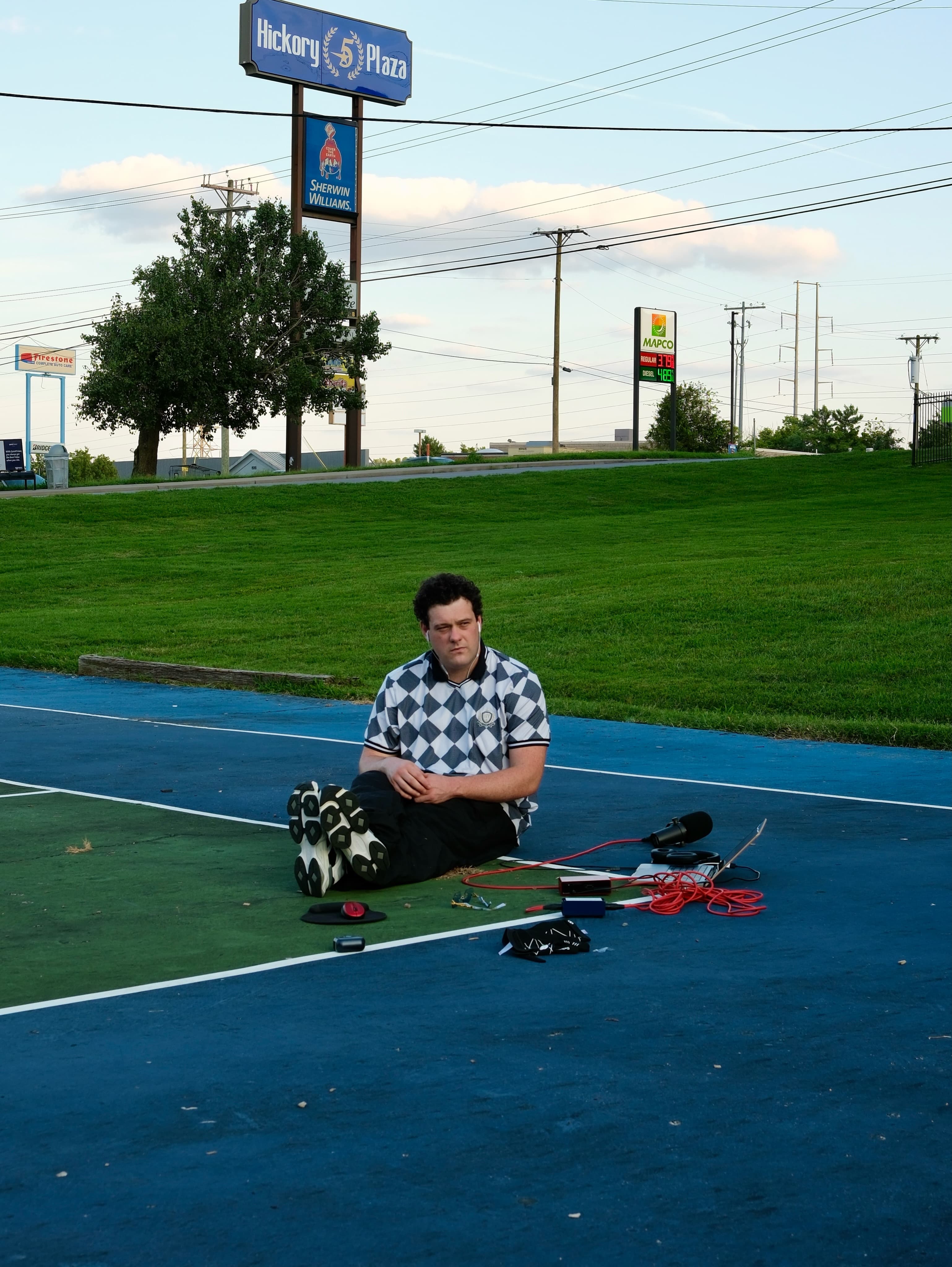 BRAVEN seated on the court in a black-and-white checkered shirt, with roadside signs behind him.
