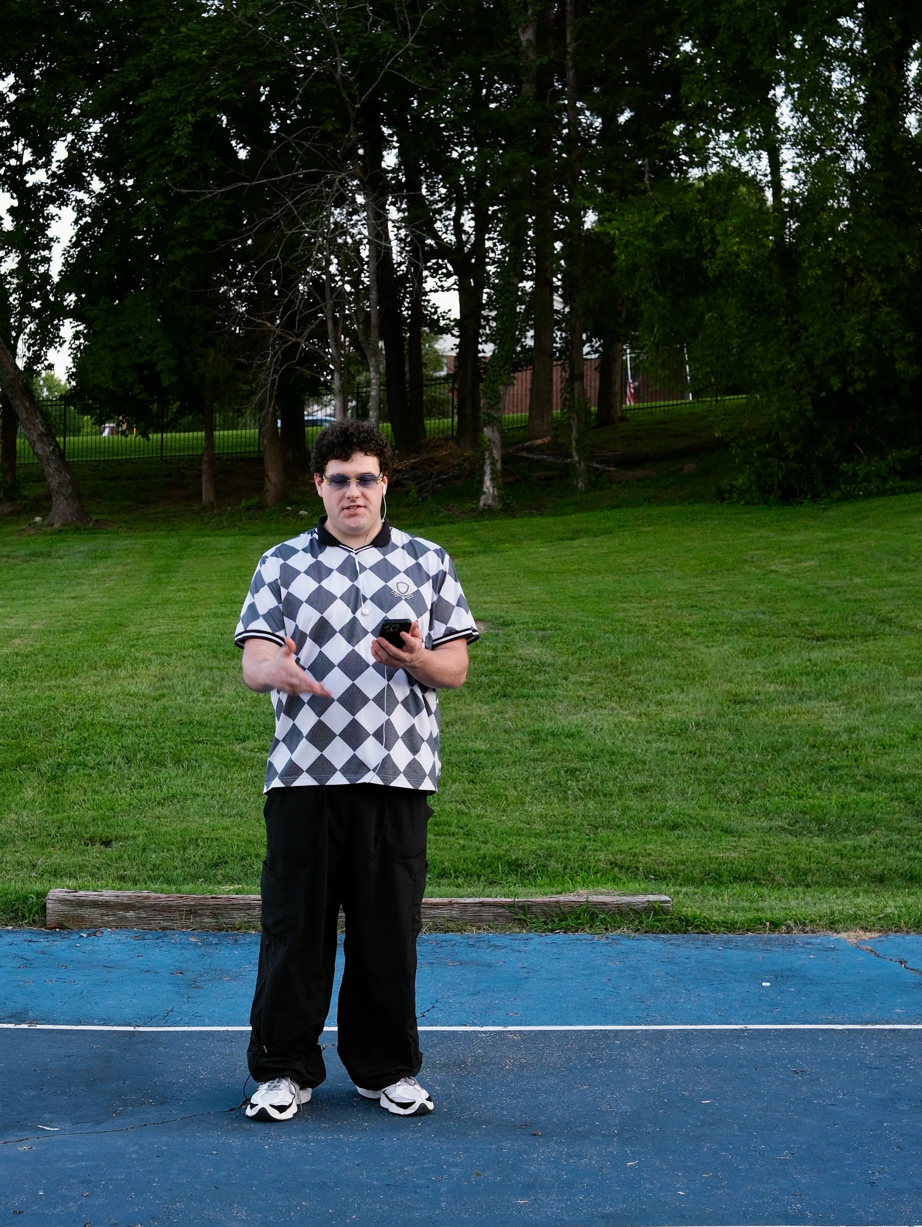 BRAVEN standing on the blue court, wearing tinted glasses and holding his phone.
